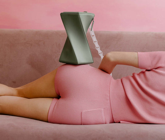 A woman lies on a pink couch with a green handbag balanced on her hip