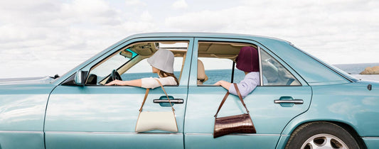 Two women sit in a light blue car with their handbags draped outside the car windows.