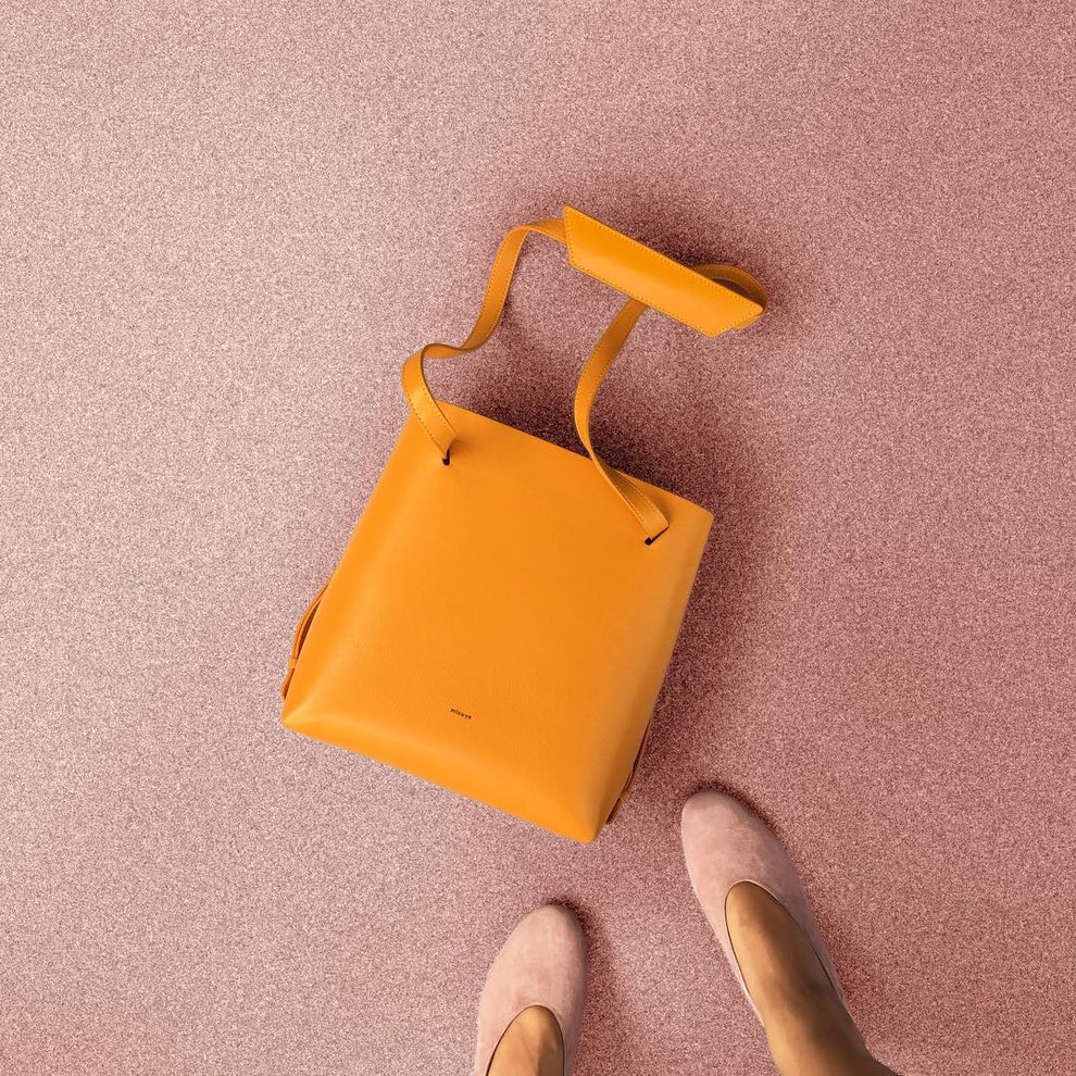 A top-down view of an orange handbag and a woman's feet wearing pink shoes. 
