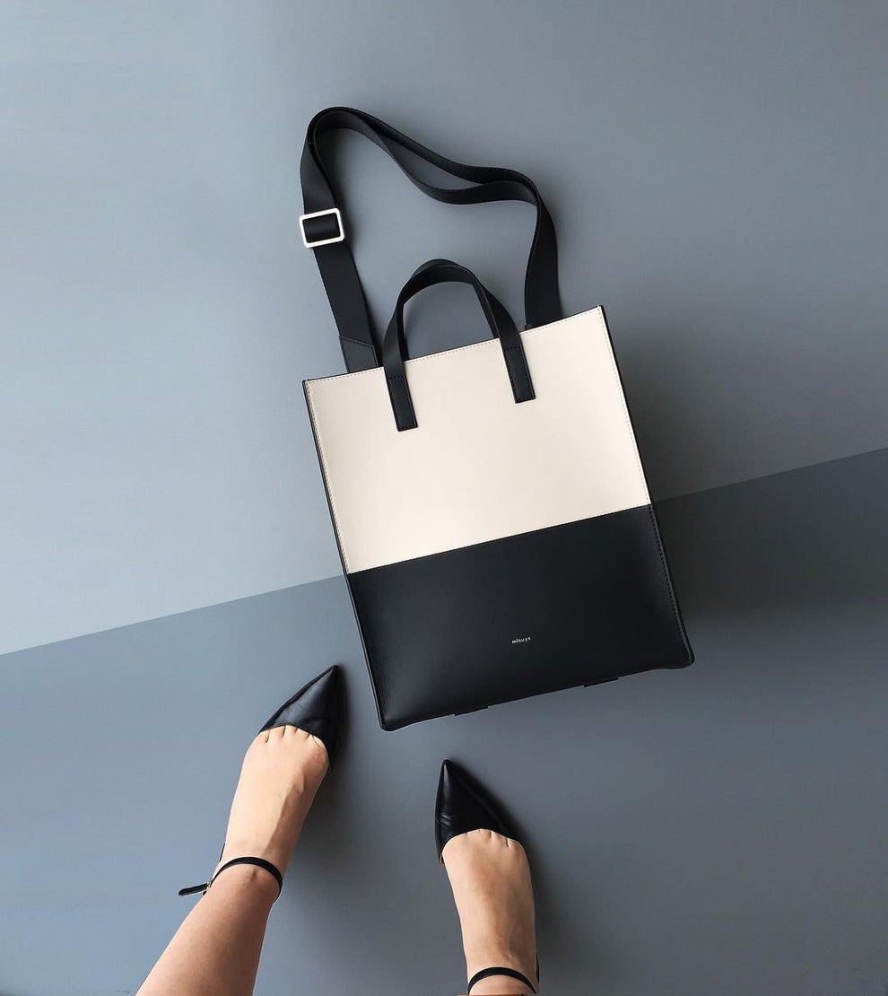 A top-down view of a black and white handbag and a woman's feet in pointed black shoes. 