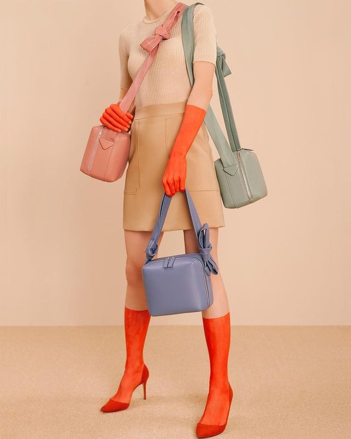 A woman wearing bright orange socks and gloves holds three handbags in different colors. 