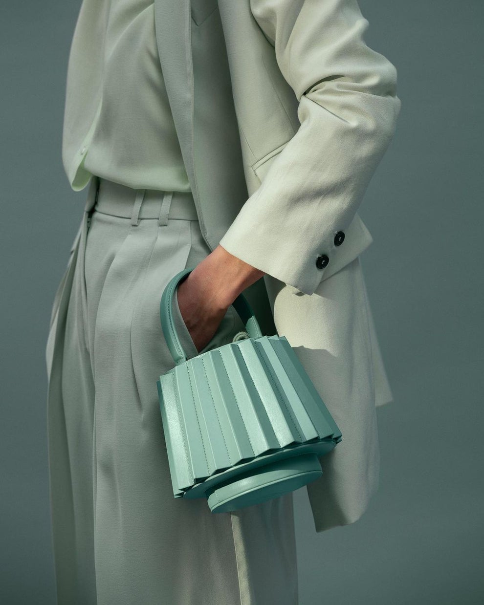 A model holds a mint green pleated lantern bag while wearing a color matched suit. 