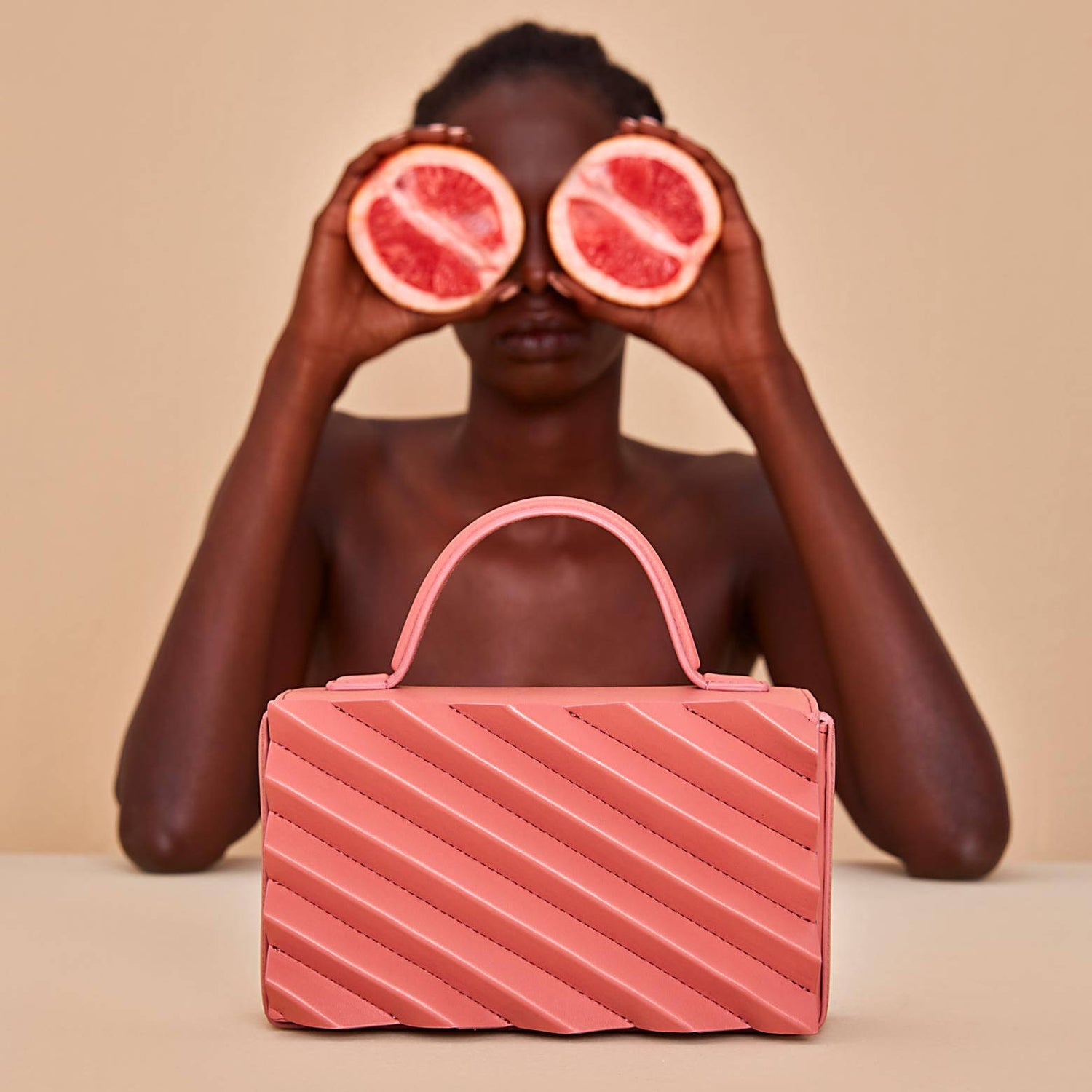 A pink handbag is displayed, with a woman in the background placing grapefruit slices playfully over her eyes. 