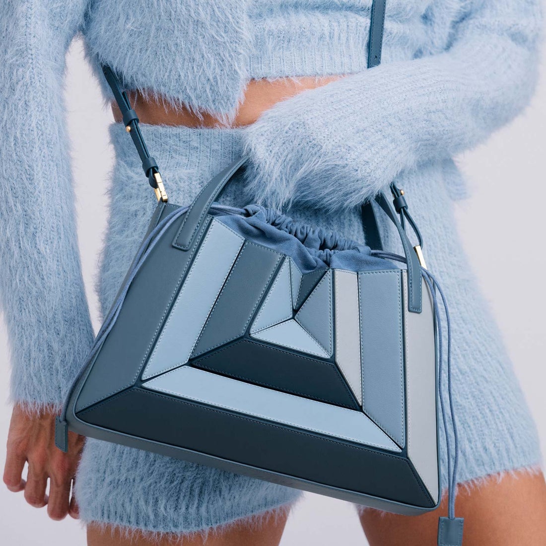 A woman models a blue handbag while wearing a fuzzy textured pale blue outfit. 